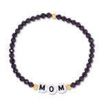 A beaded stretch bracelet featuring faceted dark purple beads, small gold spacer beads, and three white round beads spelling 'MOM' in black lettering, shown on a plain white background.