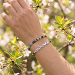 A close-up of a person's wrist wearing two beaded bracelets: one multicolor string bracelet and one white letter bead bracelet spelling 'ONE STEP'. The background is a soft-focus outdoor scene with green leaves and white blossoms.