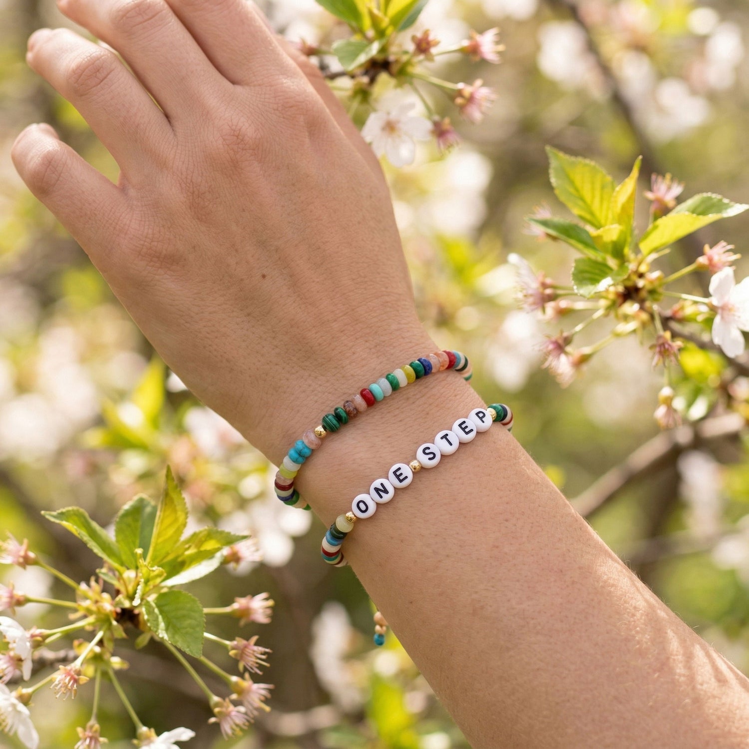 A close-up of a person's wrist wearing two beaded bracelets: one multicolor string bracelet and one white letter bead bracelet spelling 'ONE STEP'. The background is a soft-focus outdoor scene with green leaves and white blossoms.
