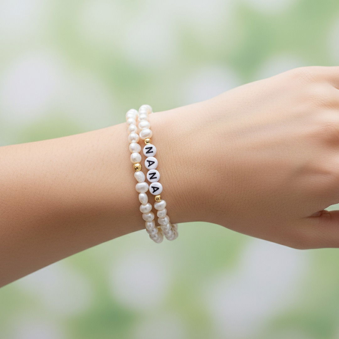 Two beaded bracelets worn on a person's wrist, featuring white freshwater pearls and white letter beads spelling 'NANA' with gold spacer beads, against a soft green and white blurred background.