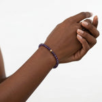 A person's wrist wearing a beaded bracelet made of dark purple beads with a small gold accent bead, shown against a plain white background.