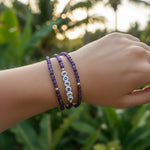 A close-up of a person's wrist wearing three beaded bracelets outdoors. Two bracelets are made of purple faceted beads with small gold spacer beads, and one bracelet features white round letter beads spelling 'SOPHIA' with gold lettering.