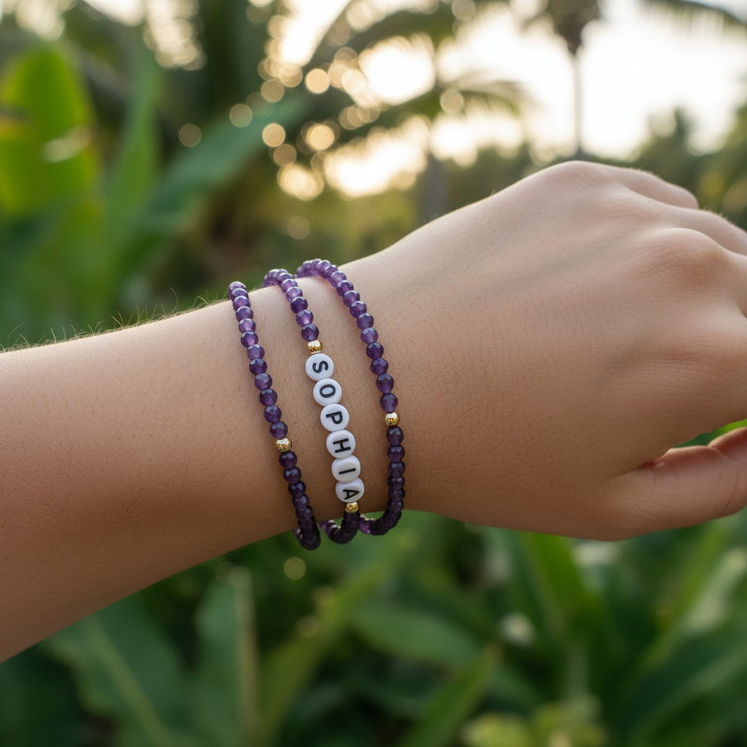 A close-up of a person's wrist wearing three beaded bracelets outdoors. Two bracelets are made of purple faceted beads with small gold spacer beads, and one bracelet features white round letter beads spelling 'SOPHIA' with gold lettering.