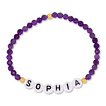 A personalized beaded stretch bracelet featuring faceted purple beads, small gold spacer beads, and white letter tiles spelling 'SOPHIA' with black lettering, shown on a plain white background.